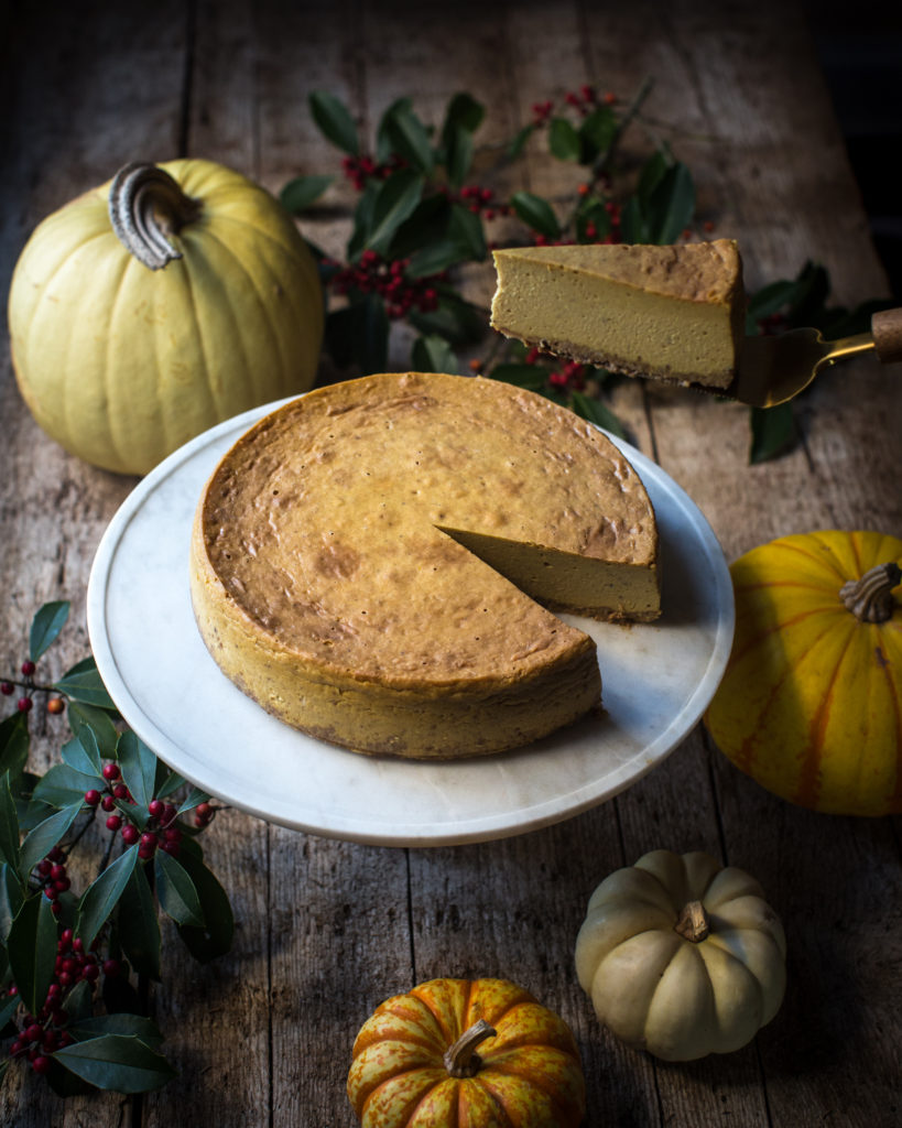 Pumpkin cheesecake on a serving tray with one slice being cut out with a background of fall gourds