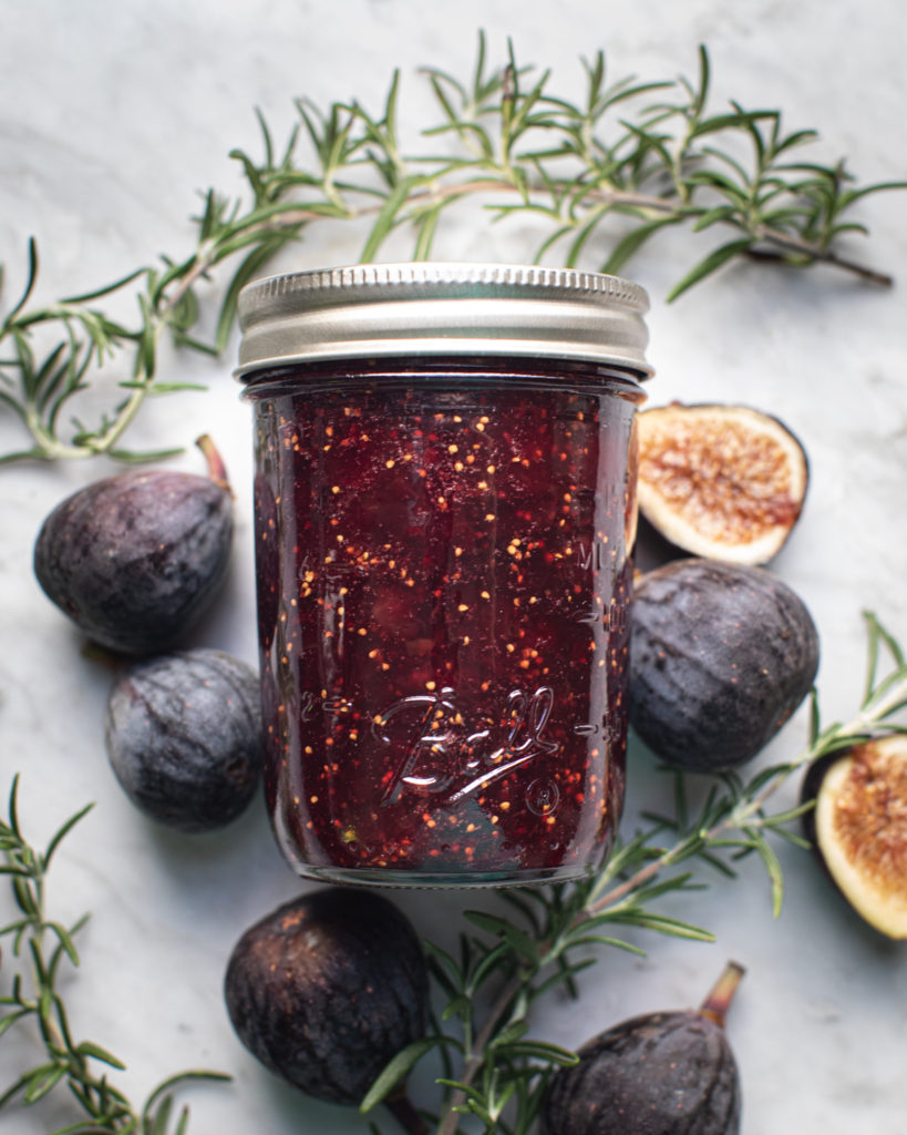 Glass jar of fig rosemary & red wine jam surrounded by figs and rosemary sprigs