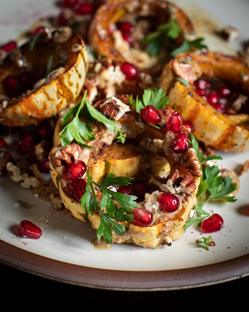 roasted delicata squash topped with pomegranate seeds, pecans, parsley and dressing on a white plate