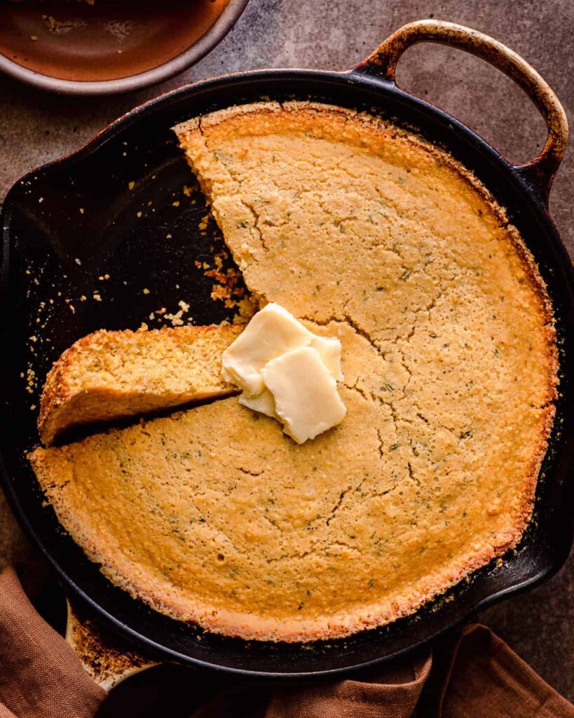 Cornbread in a cast iron skillet with a slice removed and butter on top - a must-try vegan holiday recipe!