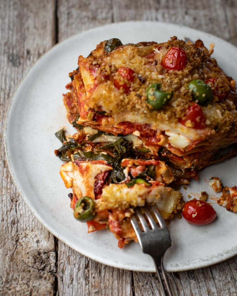 Slice of epic lasagna on a white plate with a fork taking a bite